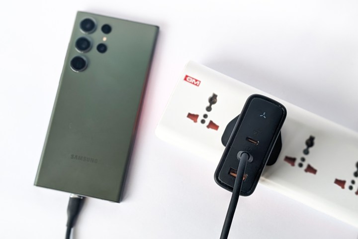 Voltme Revo 140 Gan Charger branché sur une bande d'alimentation et maintenu à côté d'un Samsung Galaxy S23 Ultra vert sur un fond blanc.