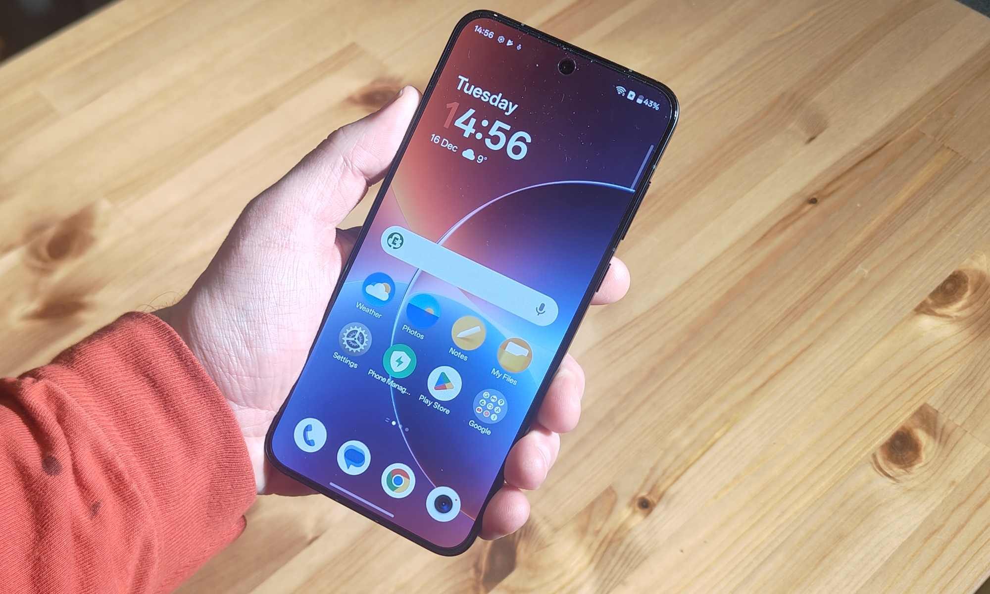 Le menu principal du OnePlus 15R, tenu dans la main d'un homme au-dessus d'une table en bois.