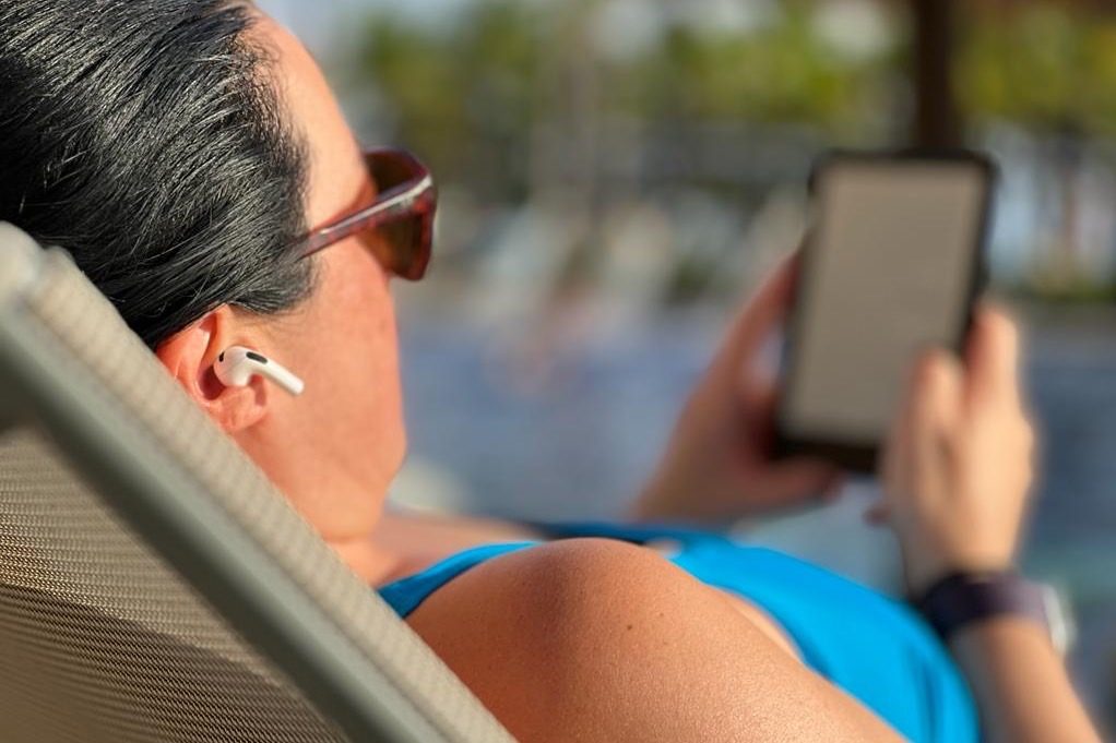 Femme au bord de la piscine avec des écouteurs Apple AirPods et une liseuse.